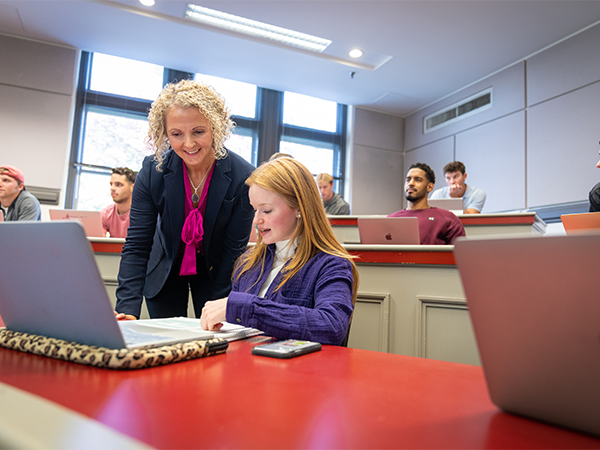 A female professor leans over the shoulder of a student looking at a packet of materials in a classroom.